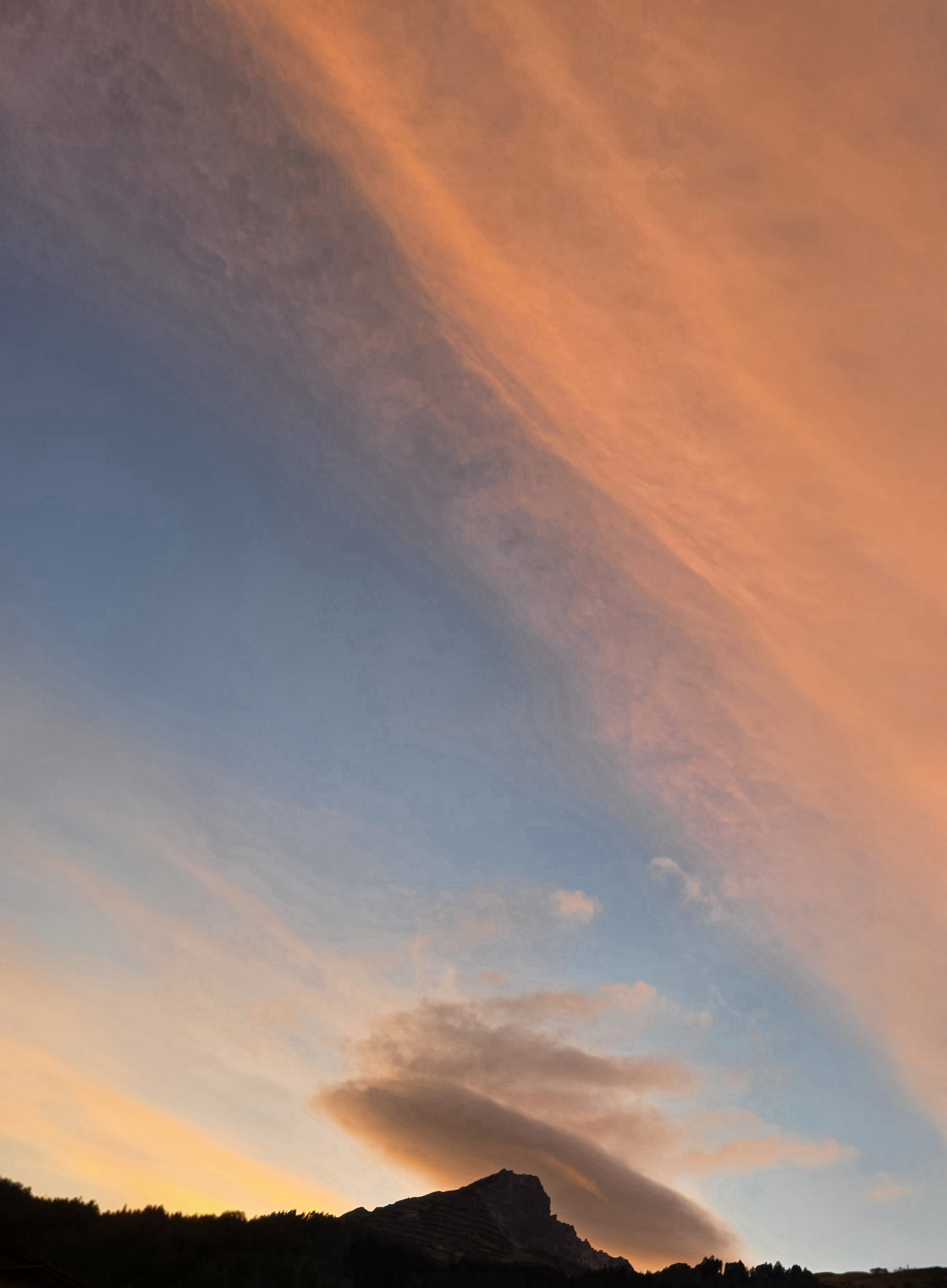 Grossflächig orange leuchtendes Wolkenband über dem dunklen Berg