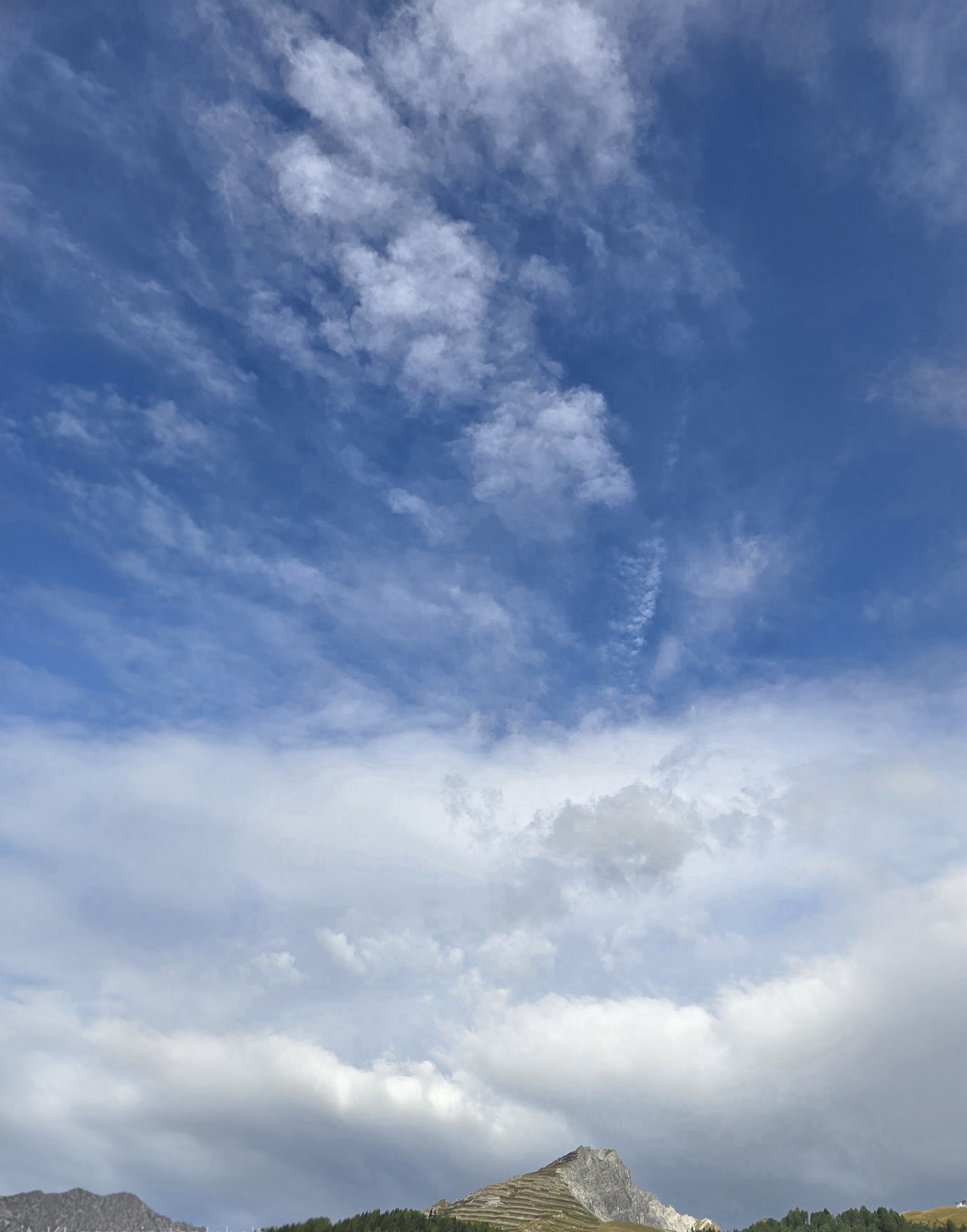Im oberen Bildteil bleibt viel blauer Himmel sichtbar. Weiter unten schieben sich grössere, helle Wolkenflächen über die Bergsilhouette.