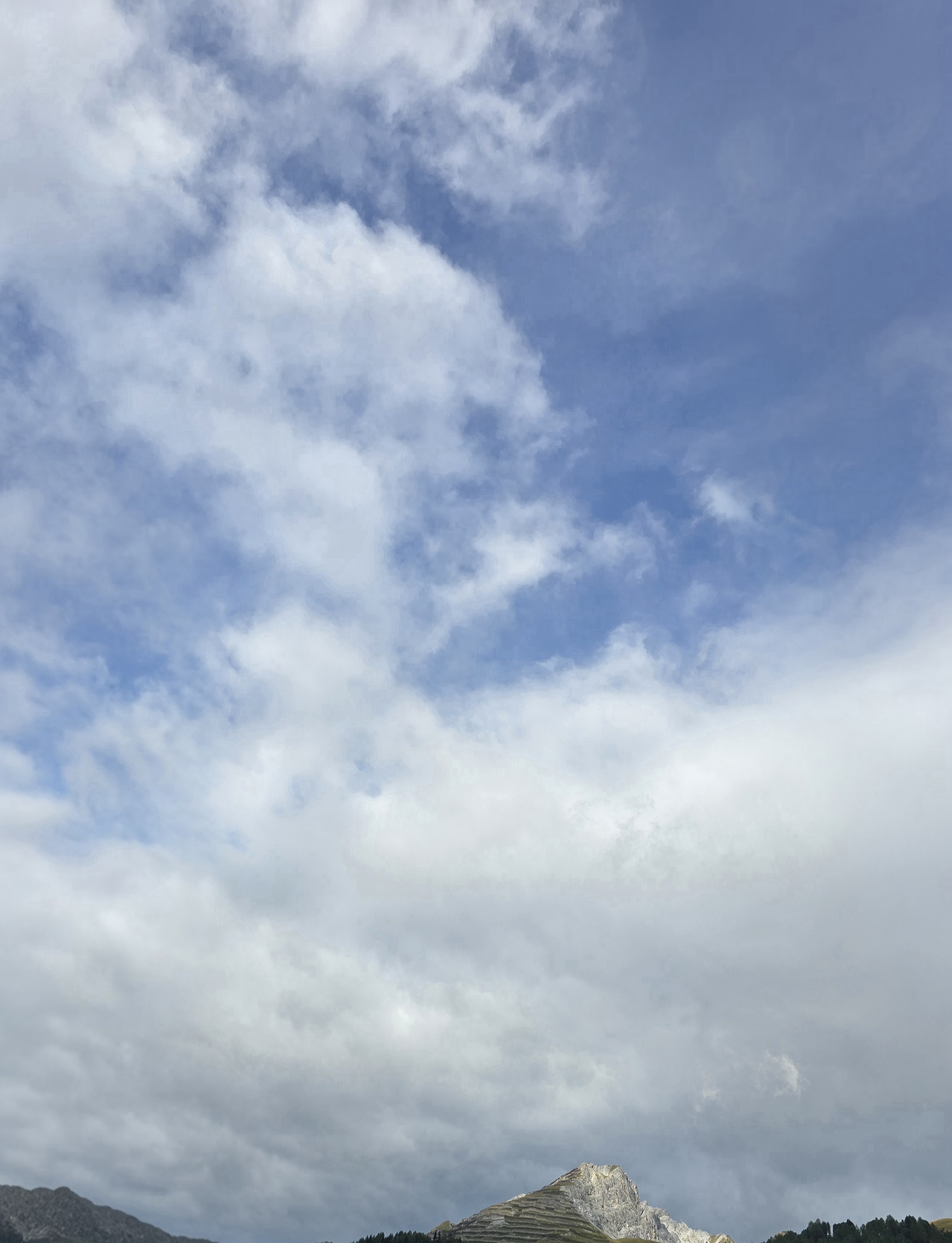 Die Wolkendecke zeigt wieder grössere blaue Öffnungen. Am Berg hebt sich eine helle Felsfläche vom dunkleren Vordergrund ab.
