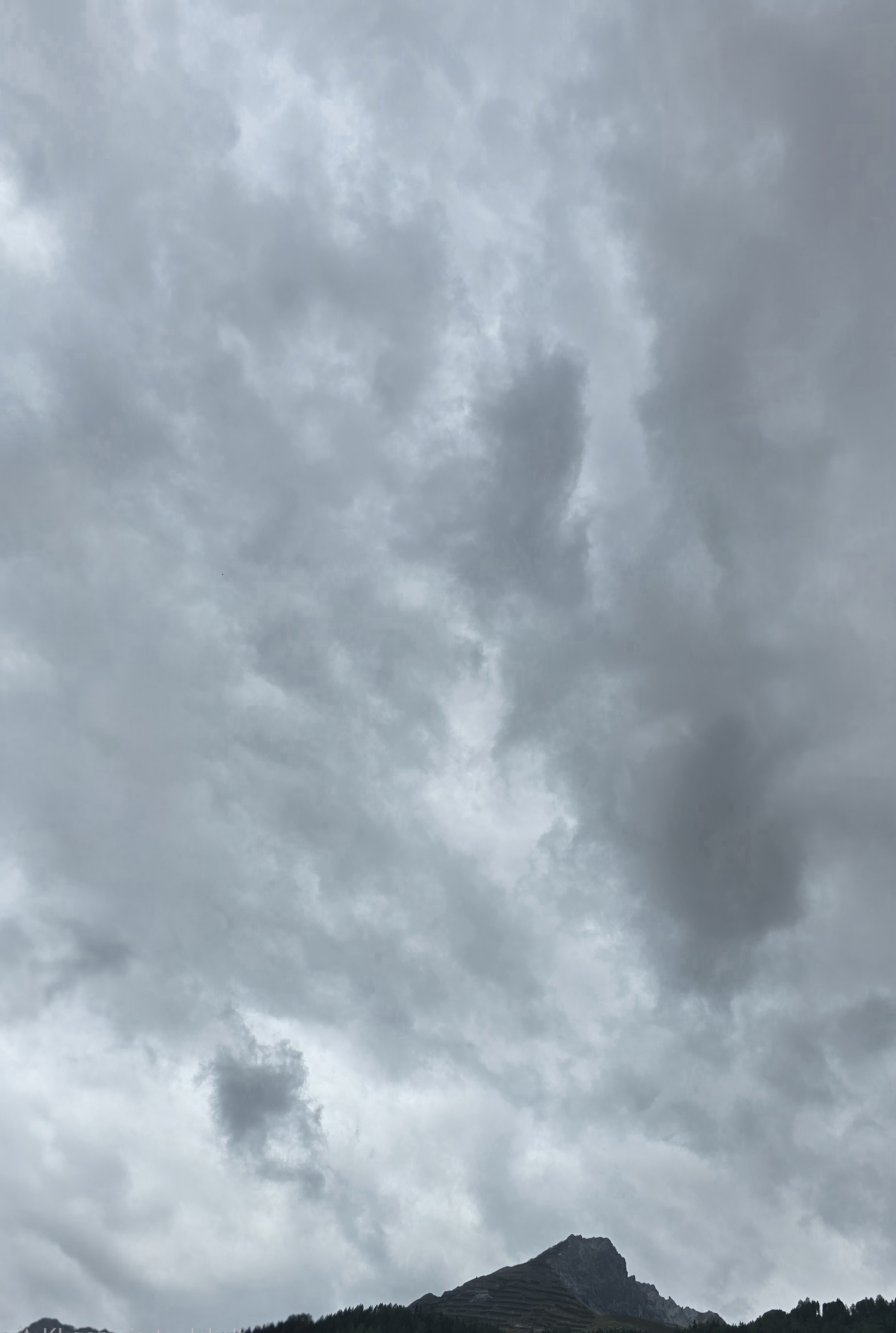 Dunklere Wolkenpartien ziehen sich durch die helle Decke. Über der Bergsilhouette bleibt kein deutliches blaues Fenster sichtbar.